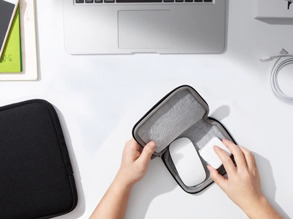 Hands placing a computer mouse into a gray protective case. Laptop, notebook, and desk accessories visible in background.