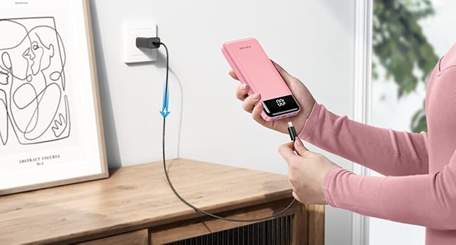 Hand holding pink smartphone with attached cable. Desk lamp and framed artwork visible on wooden surface in background.