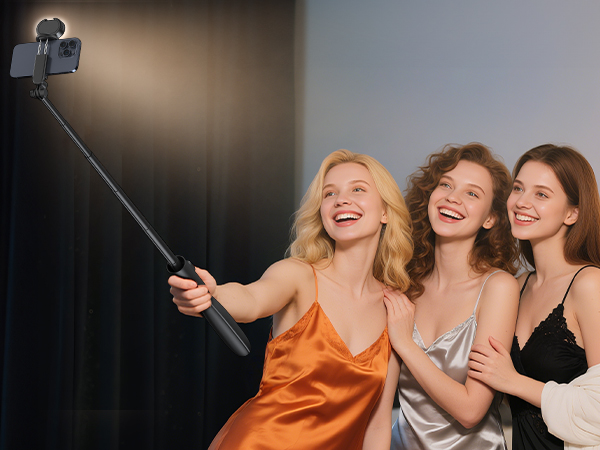 Extendable selfie stick with smartphone attached, being used to take a group photo. The device has a black handle and adjustable arm for capturing wide-angle shots.