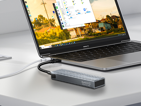 Laptop connected to a slim, dark gray USB hub or docking station. Device displays colorful desktop interface. Hub has multiple ports visible on its side.