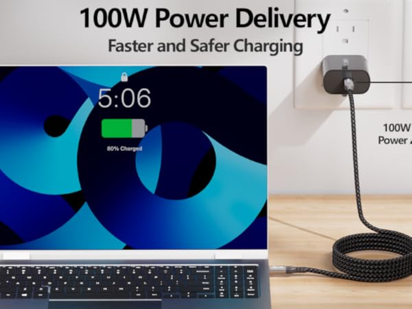 Text reads '100W Power Delivery'. Laptop displaying blue screen showing '5:06' with charging device or dock visible in foreground on wooden surface.