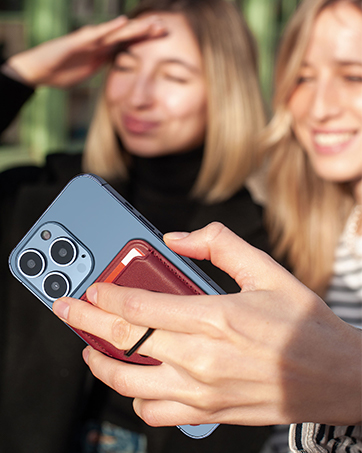 Close-up of hands holding a blue smartphone with multiple camera lenses. Two women visible in background, one shielding eyes from sun.