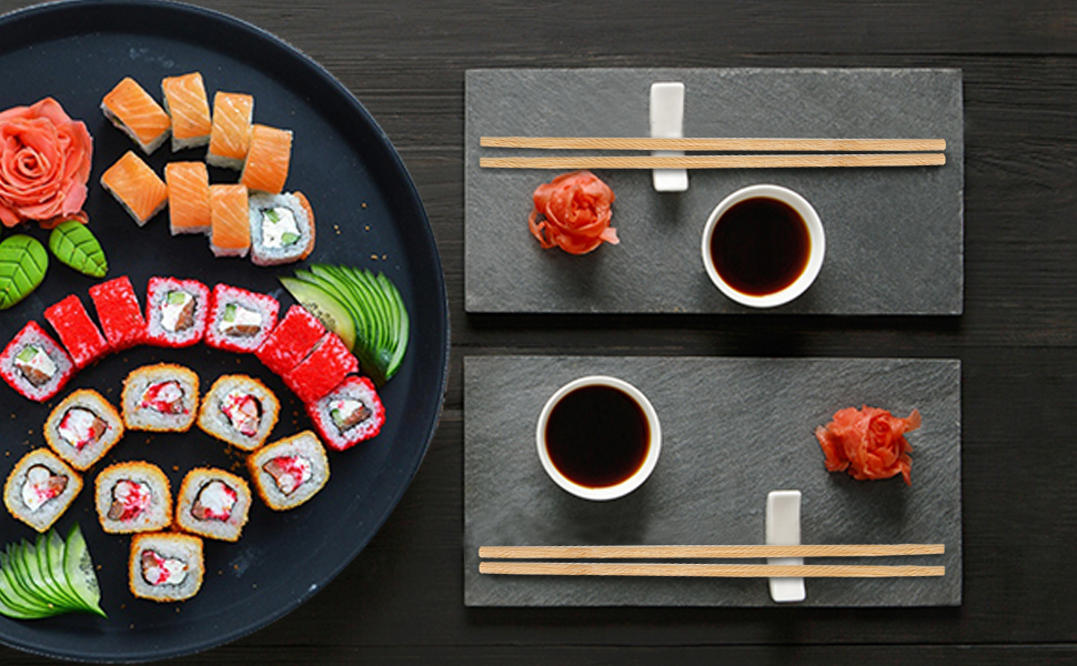 Overhead view of sushi platter arranged on black plate with rolls, nigiri, and fresh ingredients including ginger and wasabi, alongside chopsticks and two bowls of sauce on dark surface.