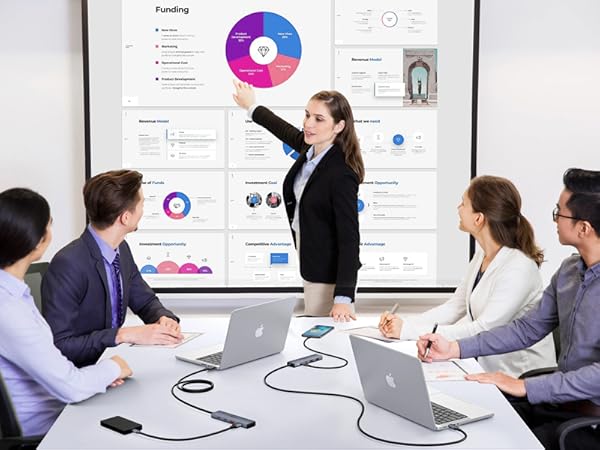 Business presentation showing colorful pie charts and data visualizations on large screen while team members view from conference table with laptops.