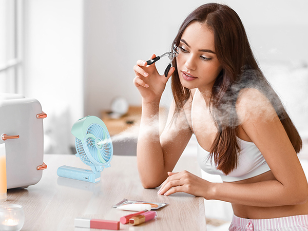 Woman using eyelash curler at vanity with cosmetics and blue fan nearby. Vaporizer emits mist in foreground.