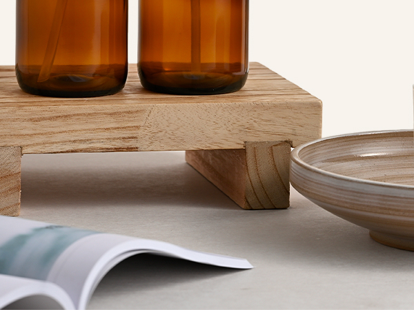 Two amber-brown glass jars with wooden base platform, displayed with neutral ceramics and fabric accessories on light background.
