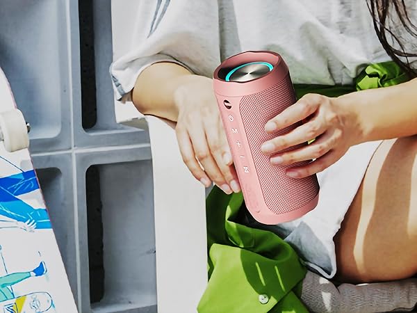Hands holding a cylindrical pink portable Bluetooth speaker with a textured surface and visible control buttons. The speaker is being carried alongside colorful beach items.