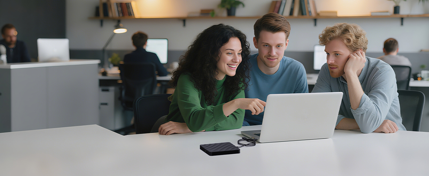 Office setting showing people collaborating while looking at a laptop screen on a desk, with office furniture and other workers visible in the background.
