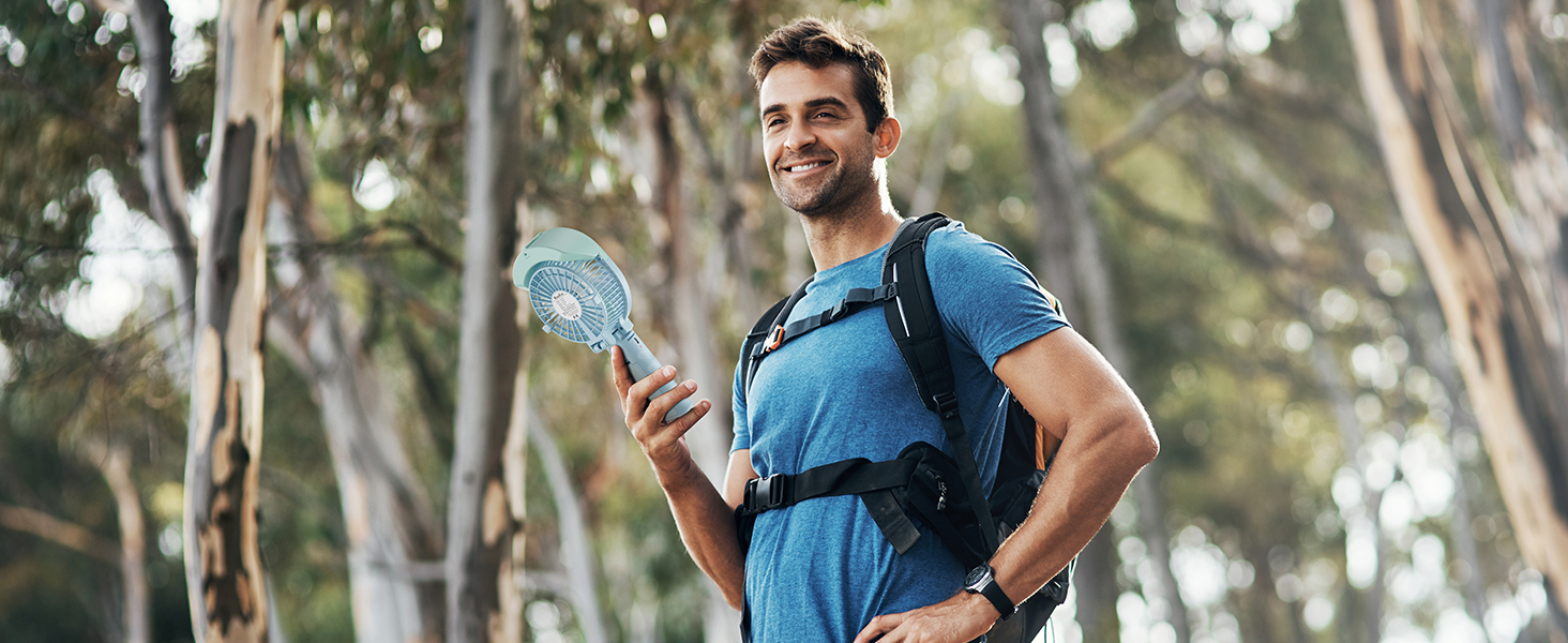 Outdoor enthusiast in blue shirt holding handheld fan in forest setting, wearing backpack with hydration system.
