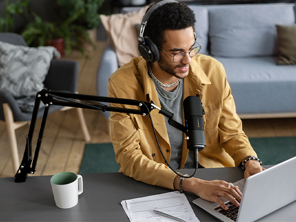 Podcasting setup with adjustable microphone arm, laptop, and over-ear headphones. Person wearing yellow jacket using equipment at a desk with coffee mug nearby.