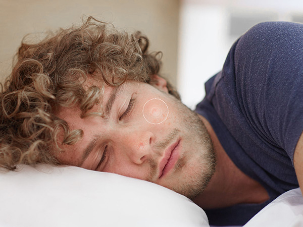 Close-up portrait of person with curly light brown hair lying down on white pillow wearing dark blue navy shirt against white background.