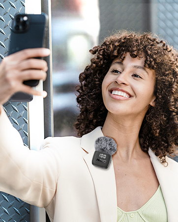 Woman with curly hair holding smartphone, smiling while taking selfie or video call, wearing beige blazer over light colored top against blurred interior background.