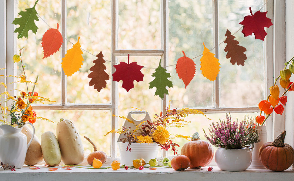 Autumn-themed windowsill display featuring orange and red maple leaf garlands, decorative pumpkins in various sizes, and potted purple flowers against a bright window backdrop.