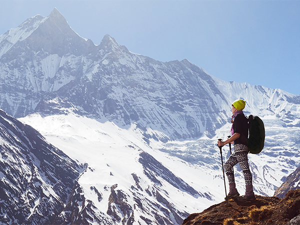 A hiker with climbing gear and backpack stands on a rocky outcrop overlooking snow-covered mountain peaks against a clear blue sky.