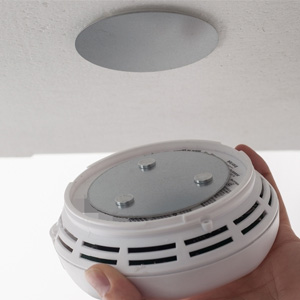 Circular smoke detector or alarm device shown installed on ceiling and held in hand. White plastic housing with vented design for sensor access.