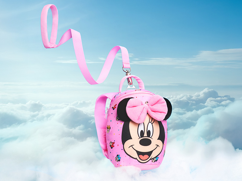 Pink children's backpack featuring Minnie Mouse design with large bow, adjustable shoulder strap, and colorful flower accents. Displayed against cloudy sky background.