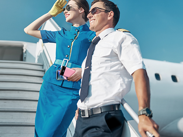 Flight attendant in blue uniform and pilot in white shirt standing on airplane stairs, shielding eyes from sun.