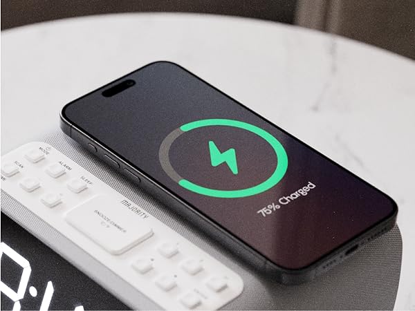 Smartphone showing 75 percent charging status on digital alarm clock with two rings bedside table