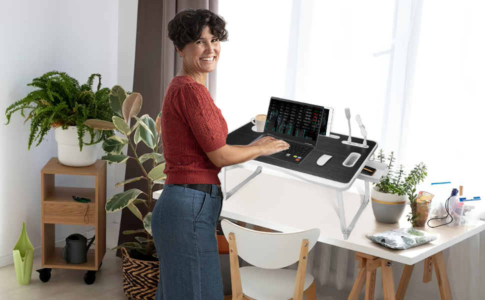 Person using a standing desk with laptop, surrounded by plants. Desk has adjustable height feature for ergonomic work positioning.
