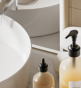 Bathroom corner with white sink, wooden cabinet, and two amber glass soap dispensers with black pumps on a white surface.