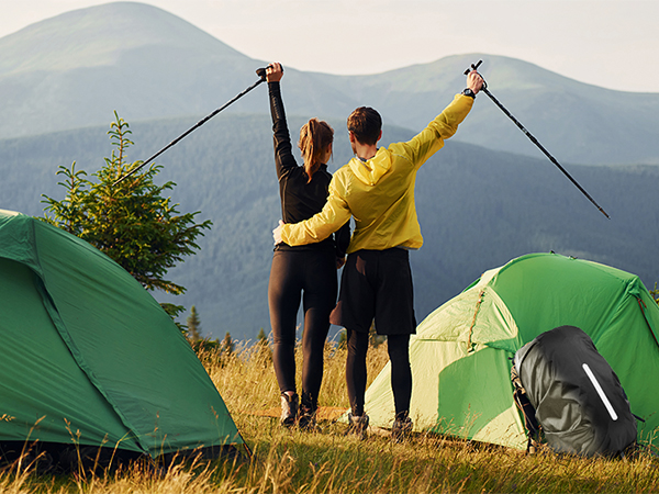 Two people celebrating on a hilltop with raised trekking poles, surrounded by green camping tents against a mountainous backdrop.