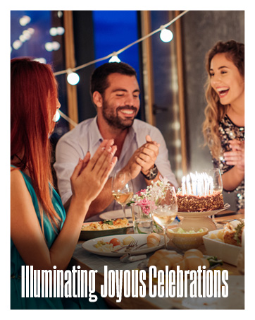 An outdoor evening lit by string lights, with three happy adults gathered around a birthday cake.
