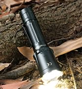 Black tactical flashlight illuminated on wooden surface against textured bark background, showing bright beam of light.