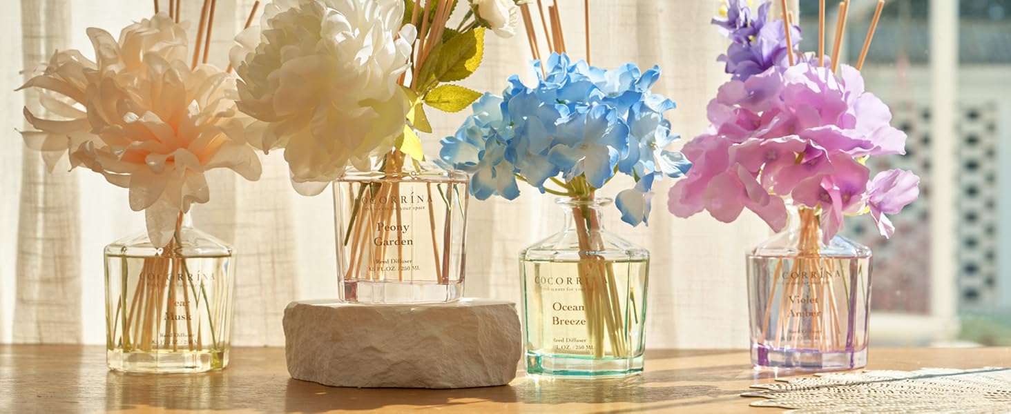 Four glass vases with colorful artificial flowers and reed diffusers, arranged on a wooden surface near a window with sheer curtains.