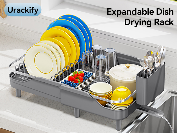 Text reads 'Urackify' and 'Expandable Dish Drying Rack'. Dark gray dish rack with expandable compartments displaying stacked blue and yellow plates, clear drinking glasses, utensil holder, and condiment containers organized in multiple sections.