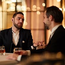 Two men in suits sitting at a bar or restaurant. One man is gesturing while speaking, creating a business or social atmosphere.
