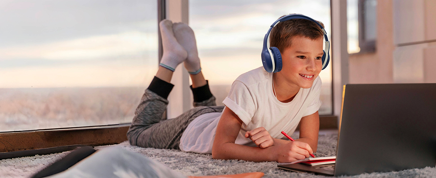 A person lying on a carpeted floor, using a laptop and wearing blue over-ear headphones. They are near a window with a view of a cloudy sky.
