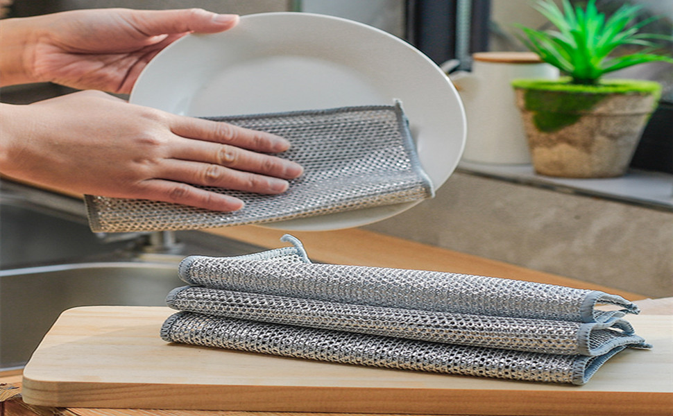Hands demonstrating use of gray microfiber cleaning cloths on a white plate. Multiple folded cloths visible on wooden surface nearby.