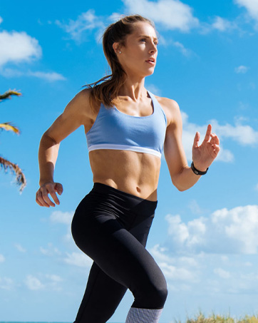 Female athlete running outdoors against blue sky and white clouds, wearing white sports bra and black leggings.