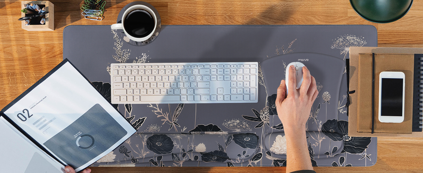 Overhead desk workspace view showing keyboard, mouse, coffee cup, smartphone, and notebook on dark desk mat with geometric pattern.