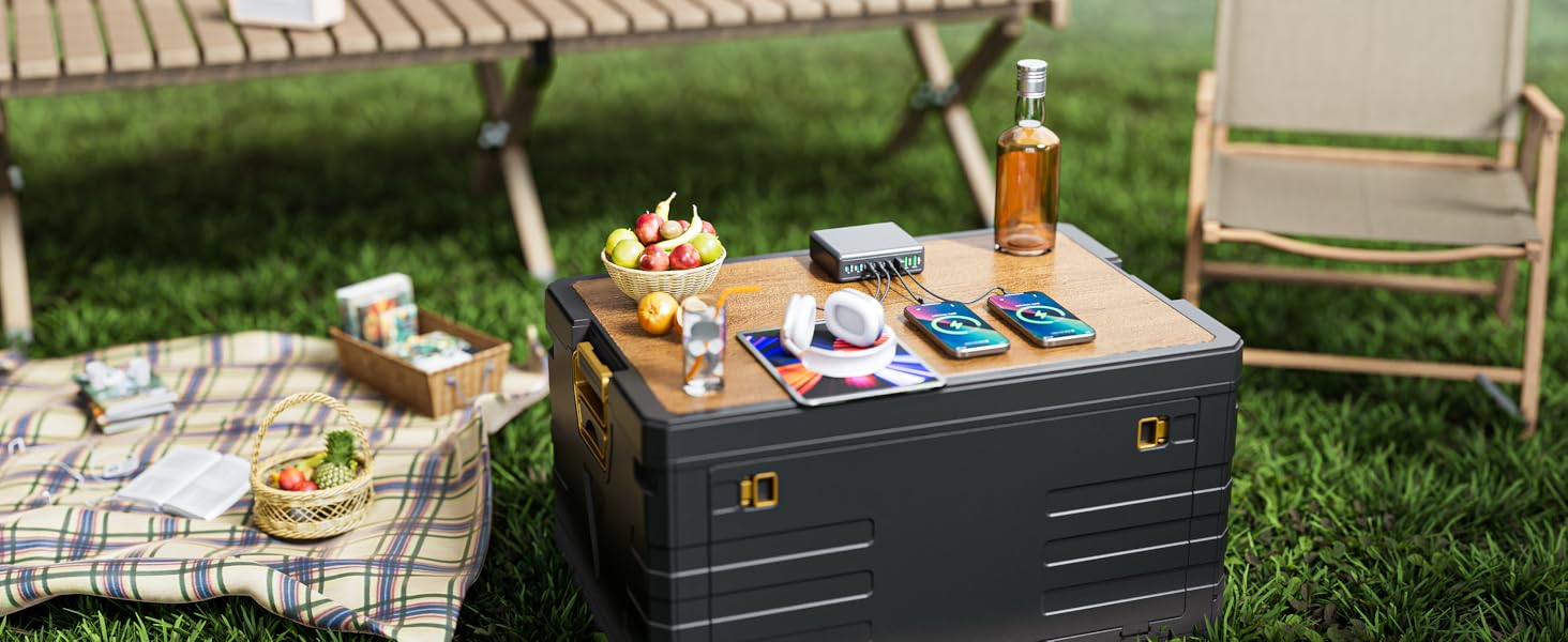 Outdoor picnic setup featuring a dark gray storage trunk used as a table, with food and drinks arranged on top, set on grass.