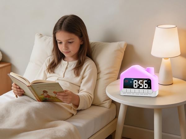 Child in bed reading book with white bedding and pillows. Purple digital alarm clock displaying 8:56 on white nightstand. Lit white table lamp beside clock.