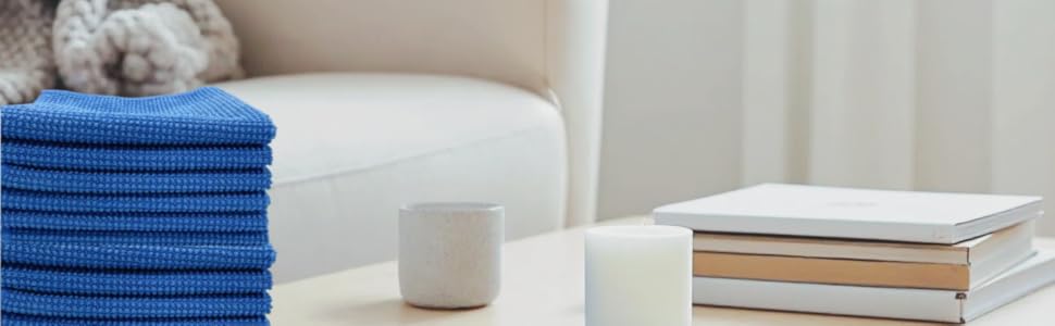 Modern living room setting with stacked blue towels, white candles, and books arranged on a light wooden coffee table near a white sofa.