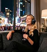 Woman wearing black clothing and headphones sitting indoors by window at night. Warm interior lighting with urban nighttime cityscape visible through window behind her. Relaxed pose suggesting audio or entertainment product usage context.