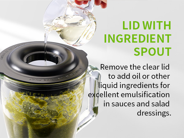 Blender jar with green contents and a black lid featuring an ingredient spout. Clear liquid pours through the spout, demonstrating its use for adding oils to sauces and dressings.