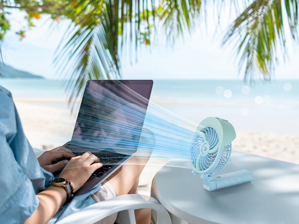 Small blue portable fan projecting air towards laptop on beach. Person typing on laptop visible in foreground, with palm fronds and ocean in background.