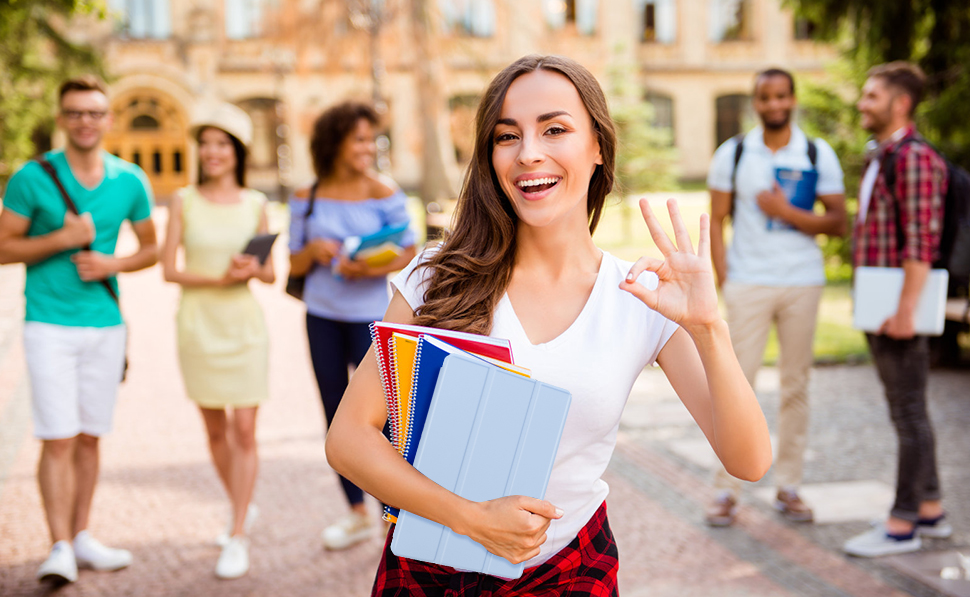 Woman holding books and smiling outdoors on campus. Other students visible in background. Sunny day with buildings in the distance.