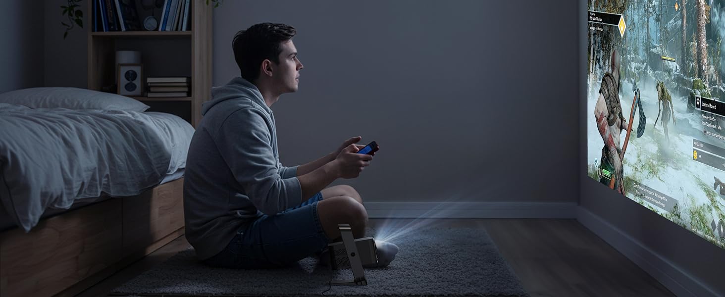 A person sits on the floor in a dimly lit room, holding a game controller while looking at a large projected image on the wall, suggesting use of a video game projector.