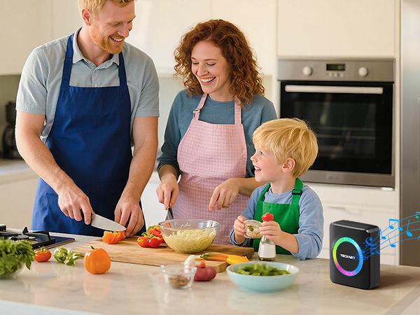 Family cooking scene in modern kitchen with small smart speaker visible. Adults and child preparing food together at counter with fresh vegetables and mixing bowls.