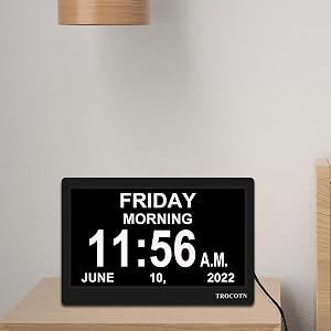 Digital clock display on a white surface, showing 'FRIDAY MORNING 11:56 A.M. JUNE 10, 2022'. A beige lampshade is visible above.
