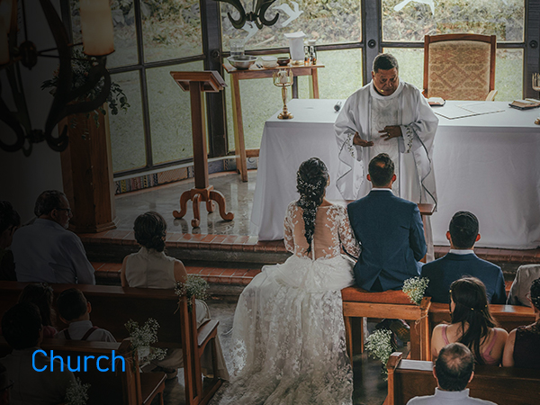 Interior of a church during a wedding ceremony. Priest stands at altar, couple and guests seated facing forward.