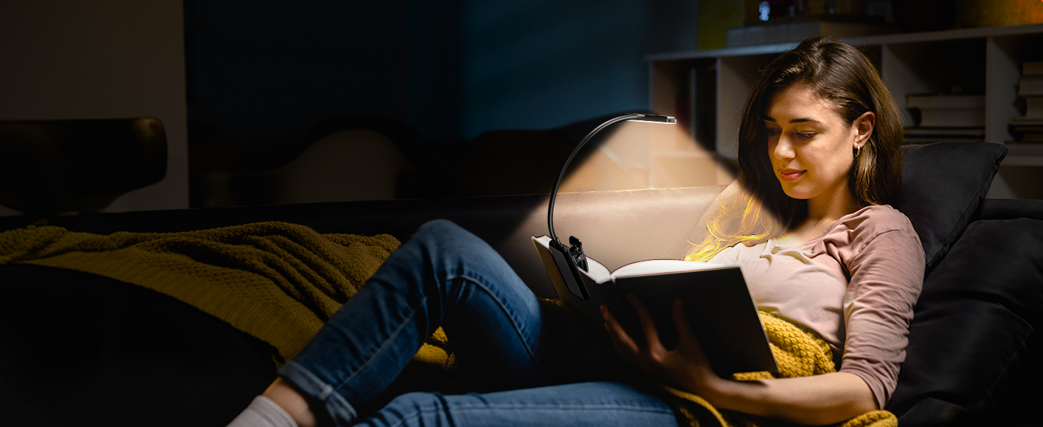A person reclining on a couch, reading a book in a dimly lit room. They are wearing casual clothing and appear absorbed in their reading.