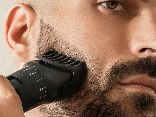 Close-up of a black electric beard trimmer in use, shaping facial hair on a man's face.
