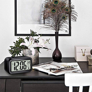 Desktop scene with digital clock, potted plant, and framed artwork. Modern, minimalist decor on dark wood surface.