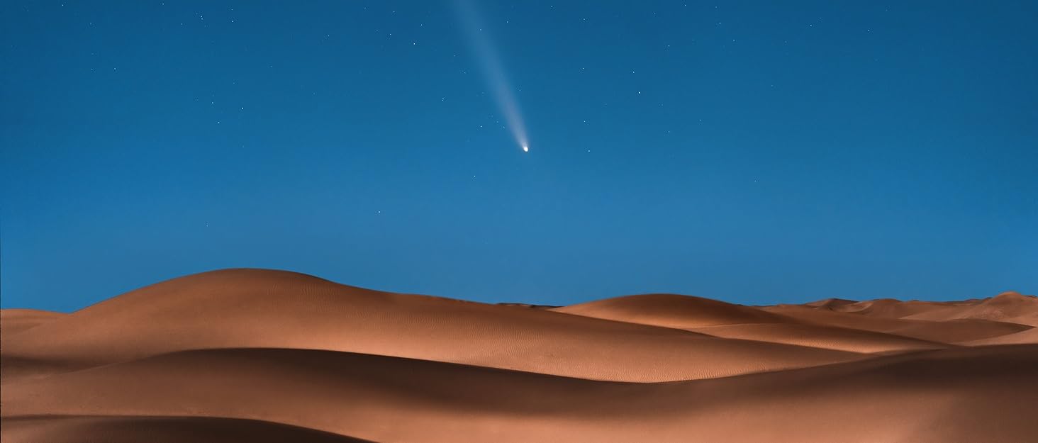 Night sky over desert sand dunes, featuring a bright comet with a long, visible tail streaking across the deep blue sky.