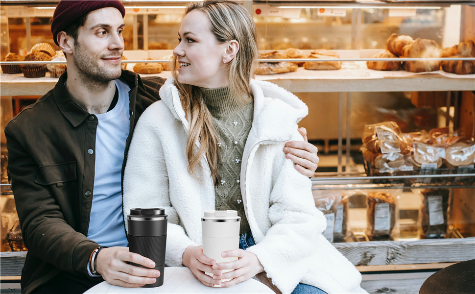 Couple smiling in a bakery or cafe, holding takeaway coffee cups. Woman wears white coat, man in dark jacket. Display of baked goods visible.